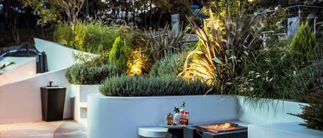 A modern outdoor seating area with a fire pit, surrounded by lush greenery and decorative lighting.