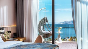 Une chambre avec un grand lit, un fauteuil suspendu, une petite table avec un vase et une vue sur la mer.