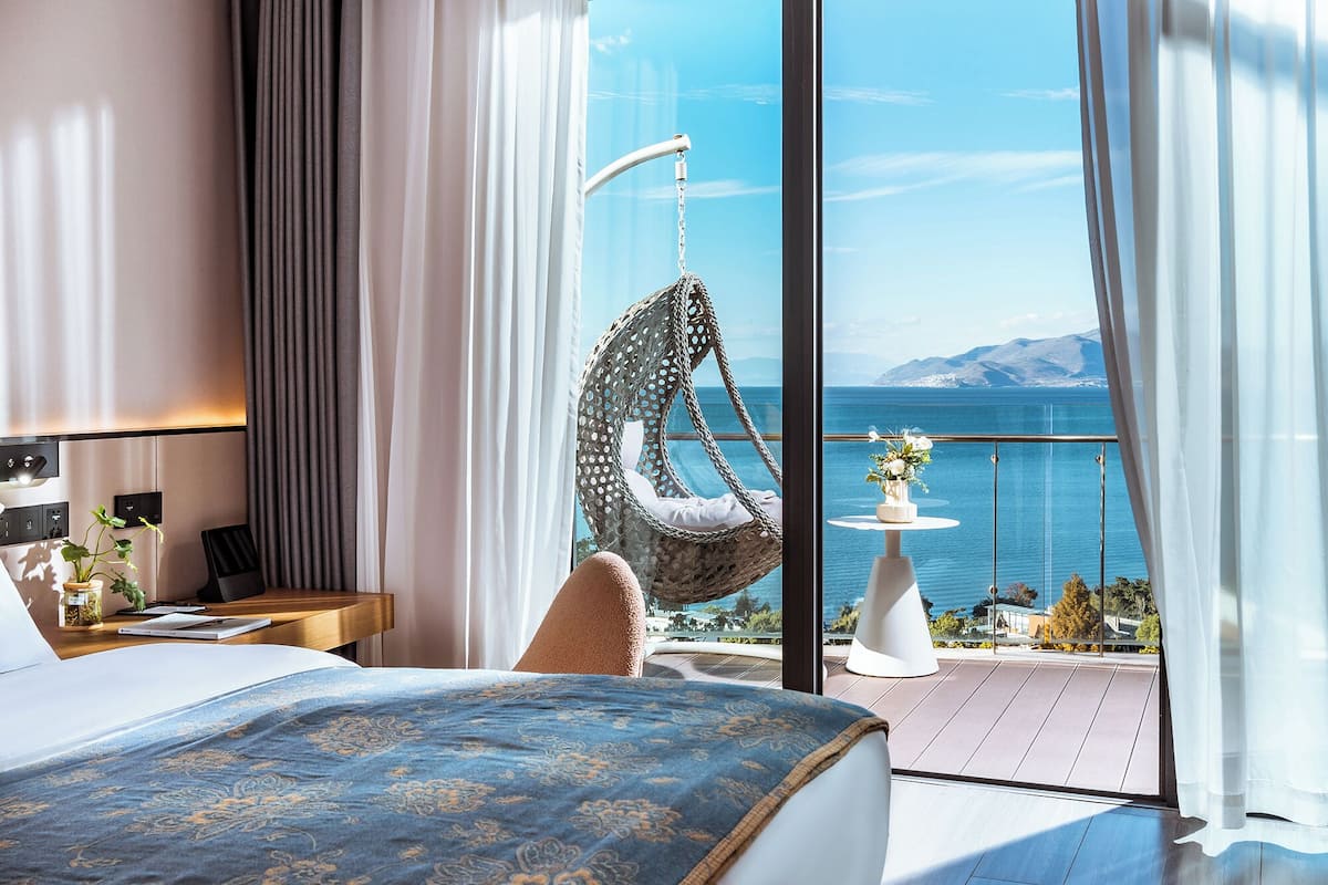 Een slaapkamer met een groot bed, een hangstoel, een klein tafeltje met een vaas en uitzicht op zee.