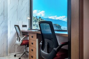 Una silla de oficina moderna con asiento rojo y respaldo negro, ubicada frente a un escritorio con acabado de madera. Una ventana muestra un paisaje montañoso.