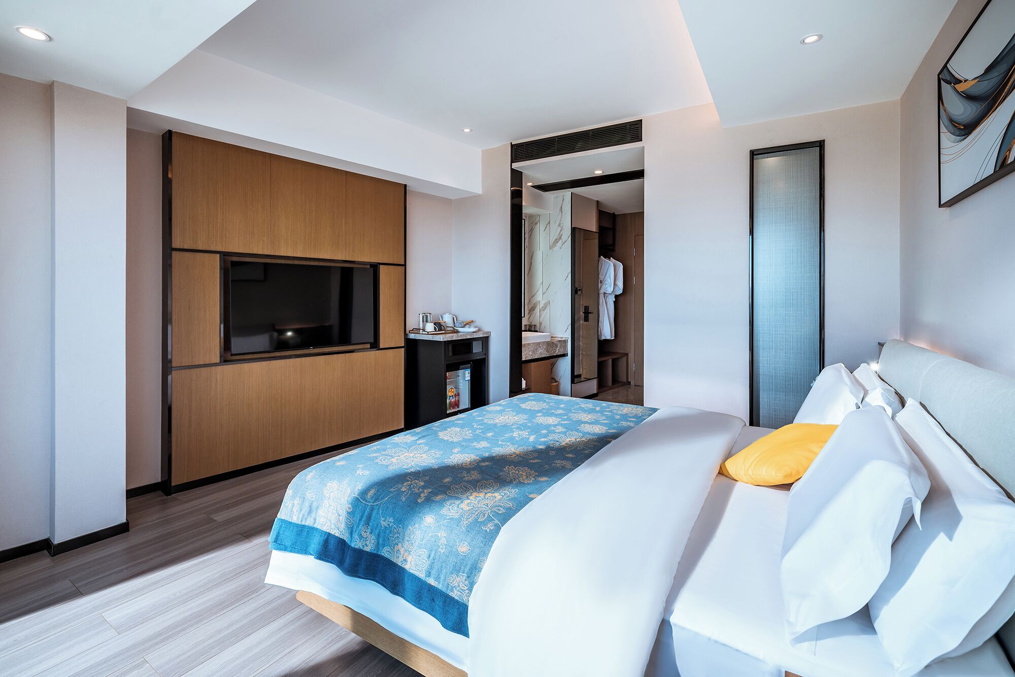A modern hotel room with a bed, a TV, a minibar, and a bathroom visible through an open door.