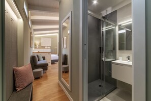 A modern hotel room with a glass shower enclosure, a sofa, a small round table, and a bed in the background.