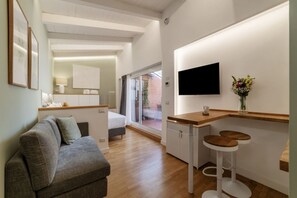 A modern, well-lit living area with a grey sofa, a wooden dining table with two stools, a flat-screen TV mounted on the wall, and a window providing a view of the outside.