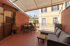 A small outdoor patio with a sofa, a table, and chairs. There is a potted plant and a hanging plant on the wall.