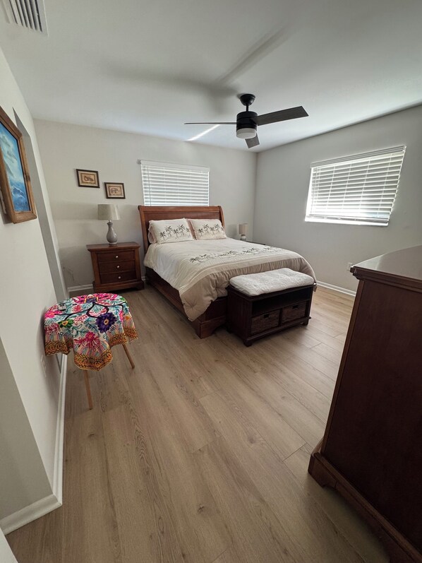 Un dormitorio con cama, mesita de noche, cómoda, ventilador de techo y dos ventanas con persianas.