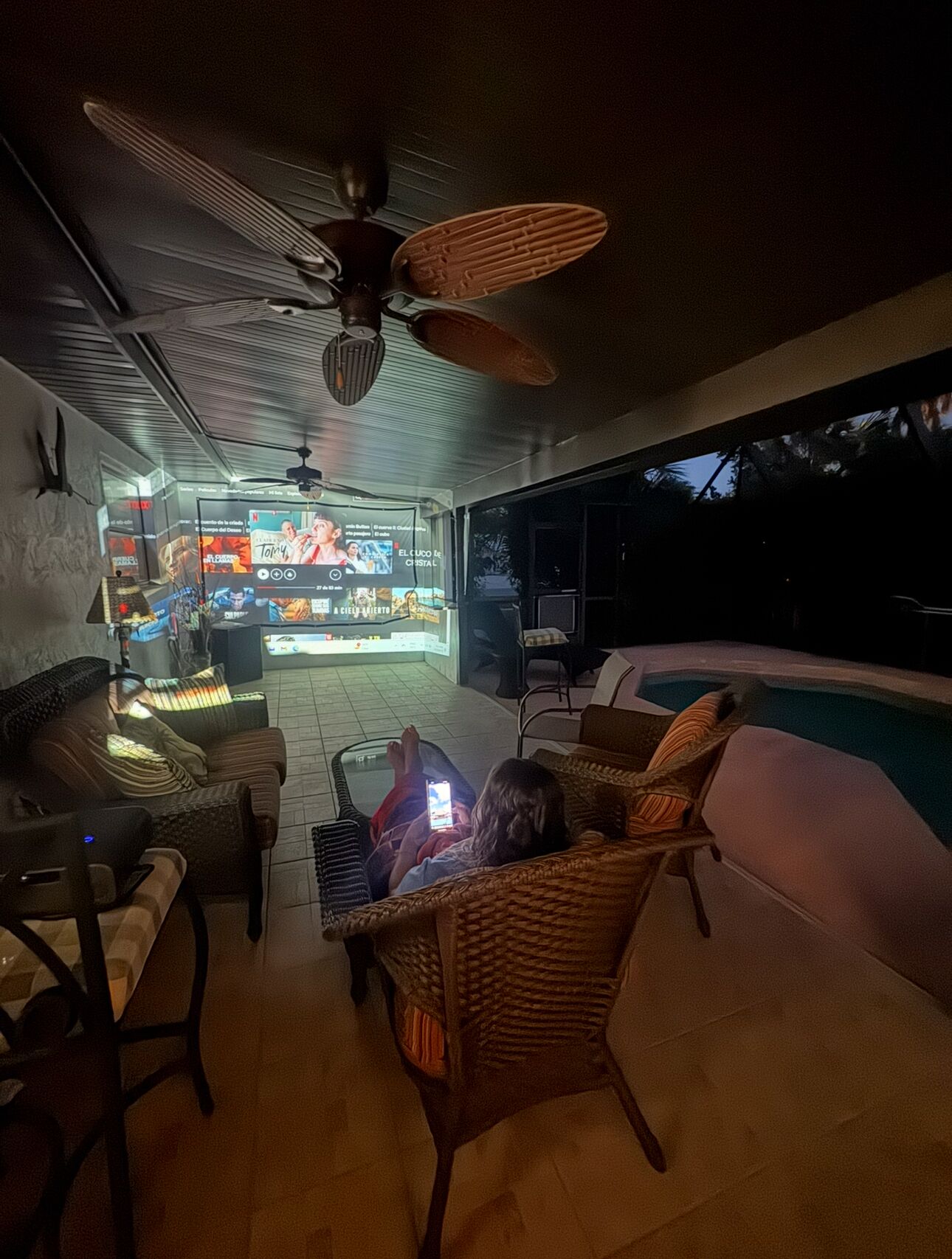 A cozy outdoor seating area with wicker furniture, a ceiling fan, and a TV screen displaying a movie.