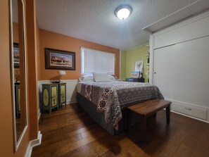Un dormitorio con cama, banco, mesita de noche, lámpara, un cuadro en la pared y una ventana con persianas.