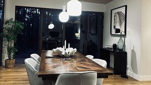 Une salle à manger moderne avec une table en bois, des chaises blanches, des luminaires suspendus et une grande fenêtre donnant sur un paysage arboré.