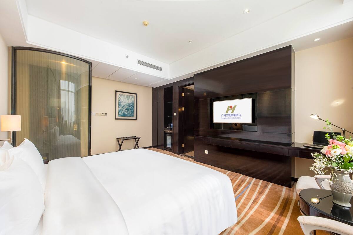 A hotel room with a large bed, a flat-screen TV mounted on the wall, a desk with a computer, and a small table with a vase of flowers.
