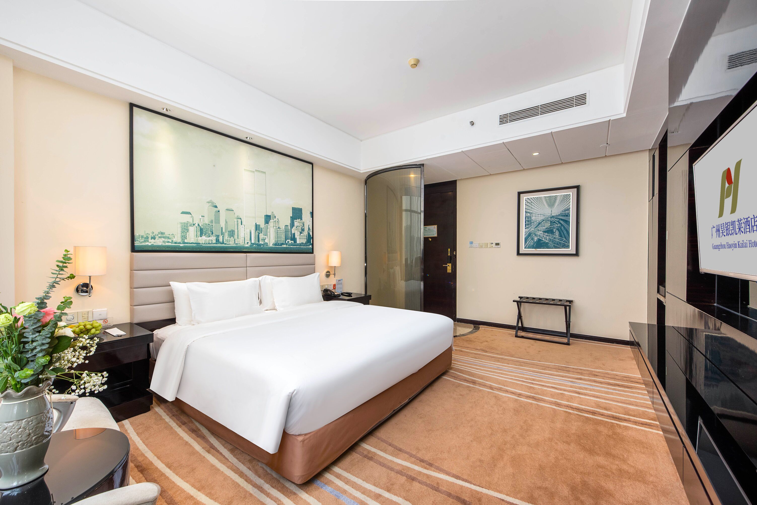 A hotel room with a large bed, a flat-screen TV, a desk with a chair, a small table with a vase of flowers, and a large window with a cityscape view.