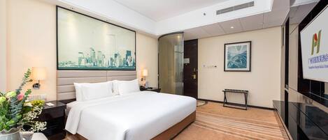 A hotel room with a large bed, a flat-screen TV, a desk with a chair, a small table with a vase of flowers, and a large window with a cityscape view.