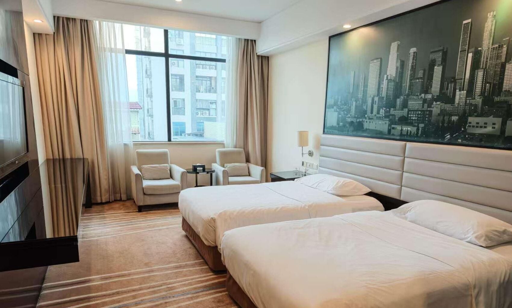 Una habitación de hotel con dos camas, un ventanal con cortinas, un cuadro de un paisaje urbano y un televisor a la izquierda.