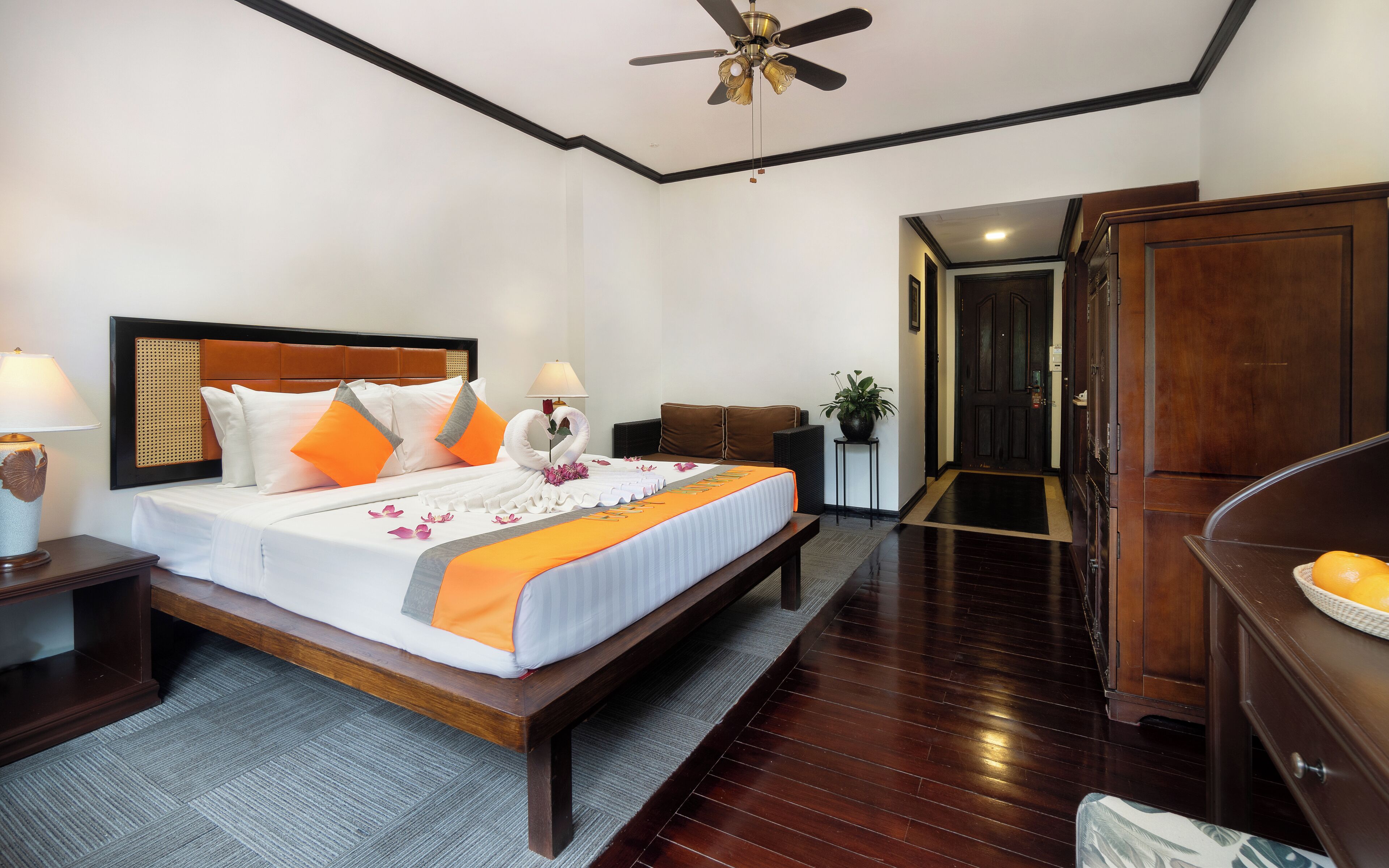 A hotel room with a bed, bedside tables, a ceiling fan, a desk, a chair, a potted plant, and a wardrobe.