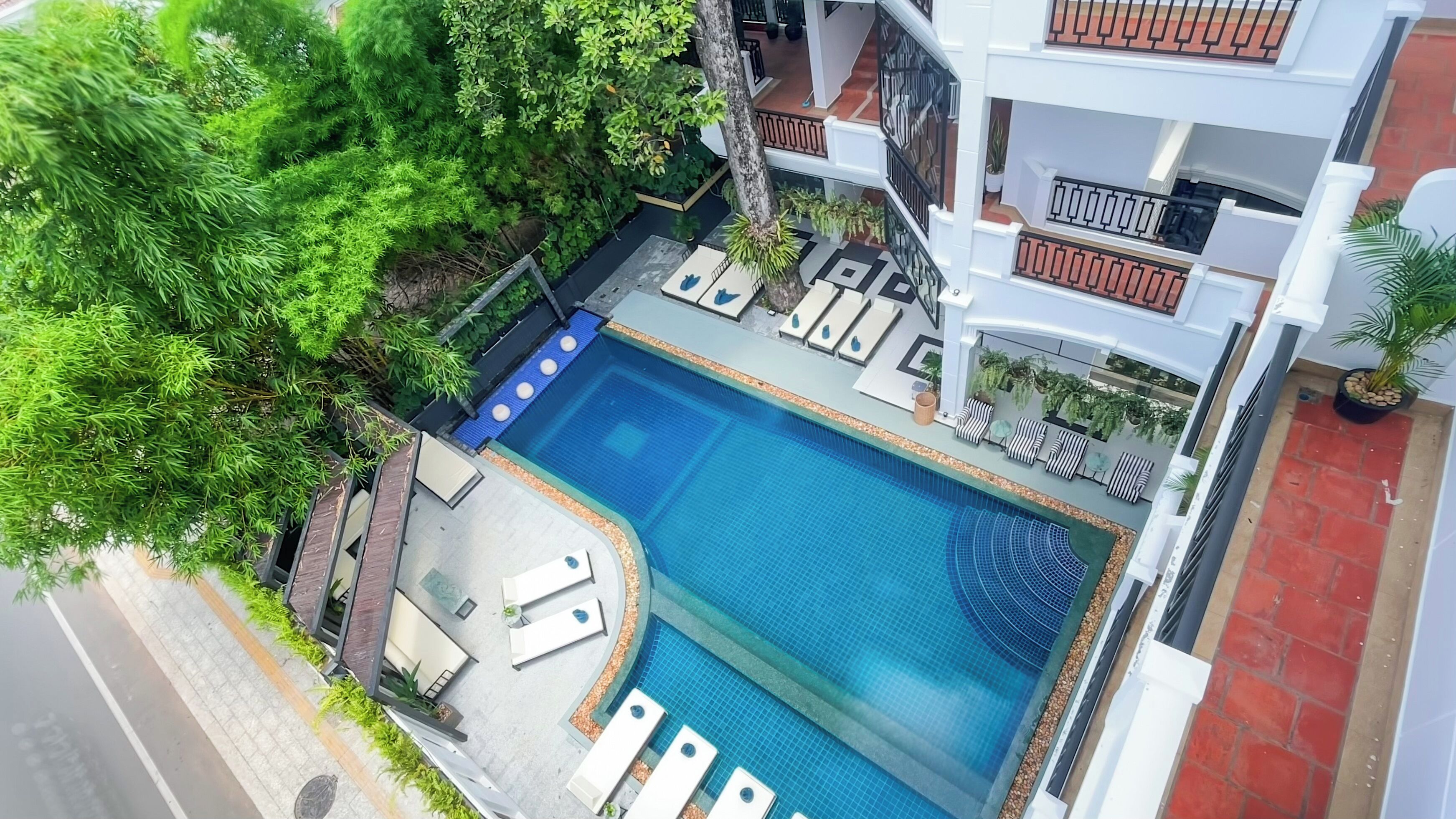 Une piscine sur le toit, dotée de chaises longues et d’un petit espace de détente, entourée de verdure et bordée d’un bâtiment blanc.