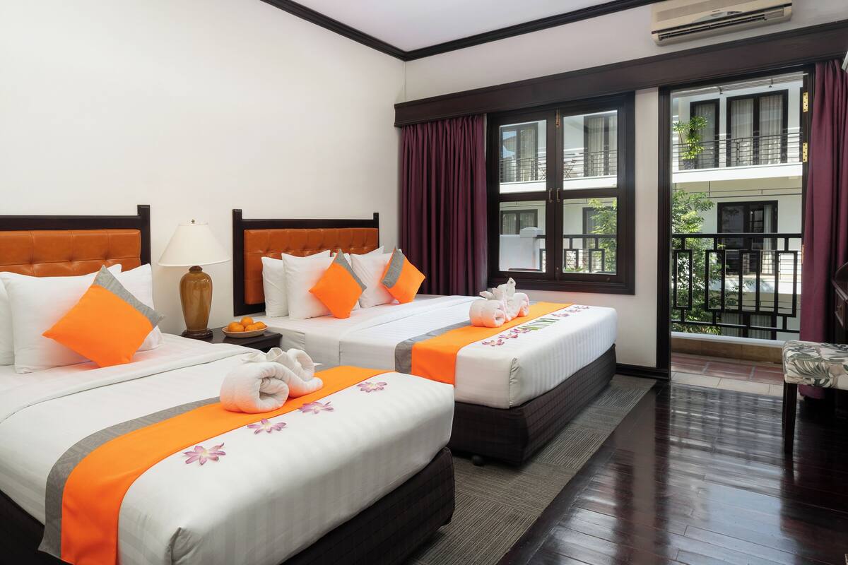 Habitación de hotel con dos camas, cada una con ropa blanca y detalles naranja, una mesita de noche con lámpara, y vistas al balcón exterior a través de amplias ventanas.