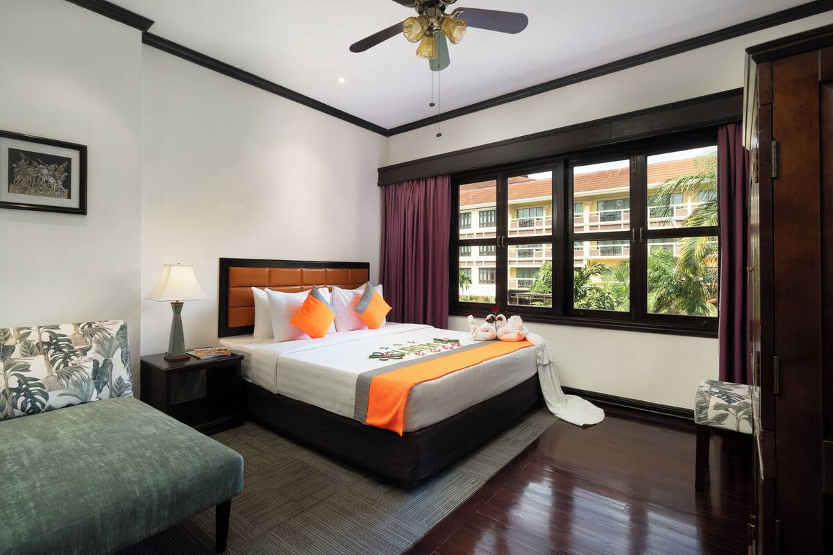 Habitación de hotel con cama, mesita de noche, lámpara, ventana con cortinas y vistas a edificios y zonas verdes.