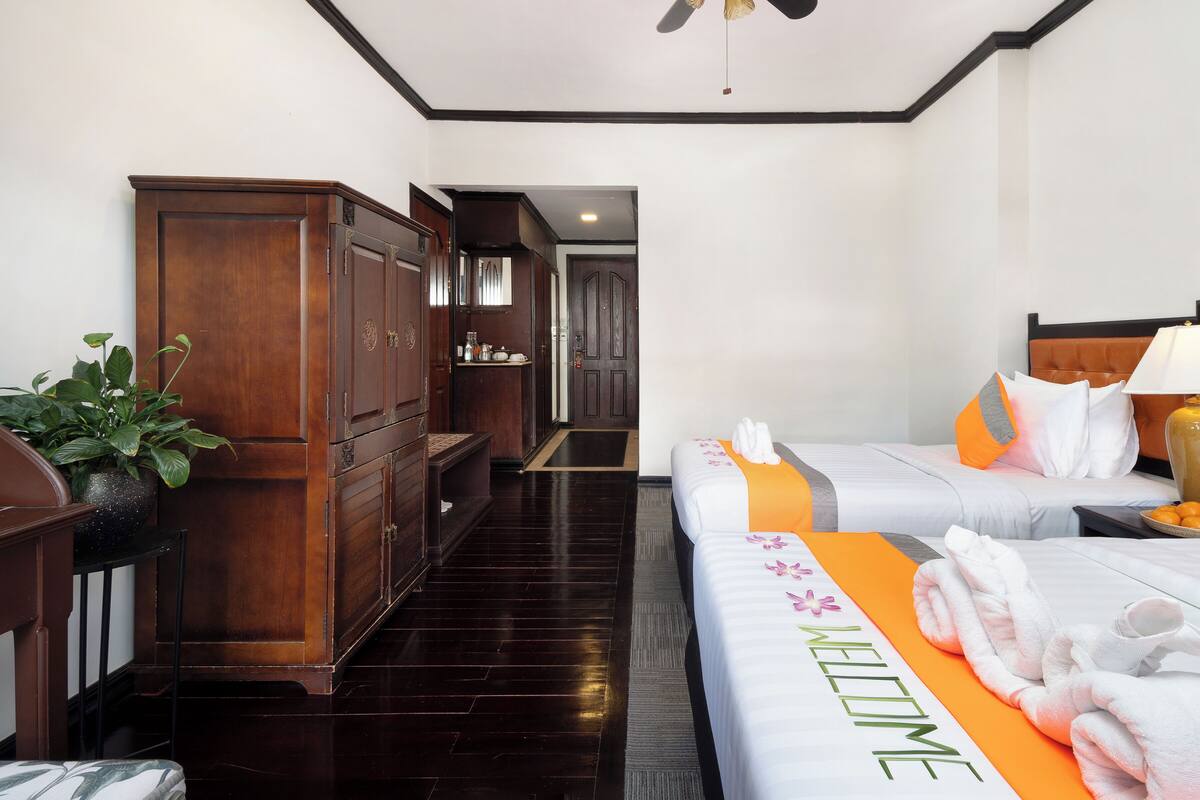 Habitación de hotel con dos camas, un armario de madera, una planta y un tapete de bienvenida.