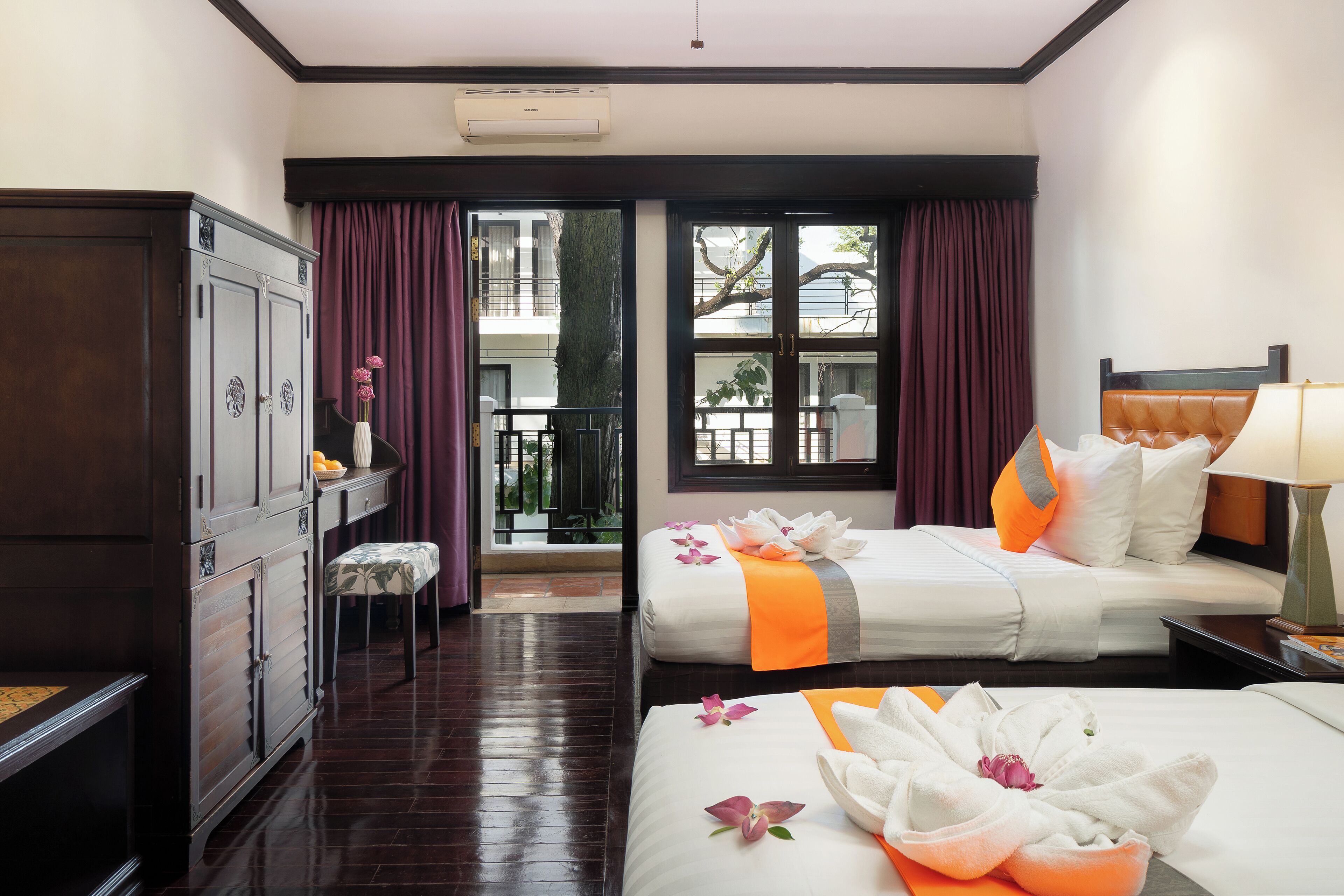 A hotel room with two beds, a wardrobe, a small table with a vase, and a view of the outside through large windows.