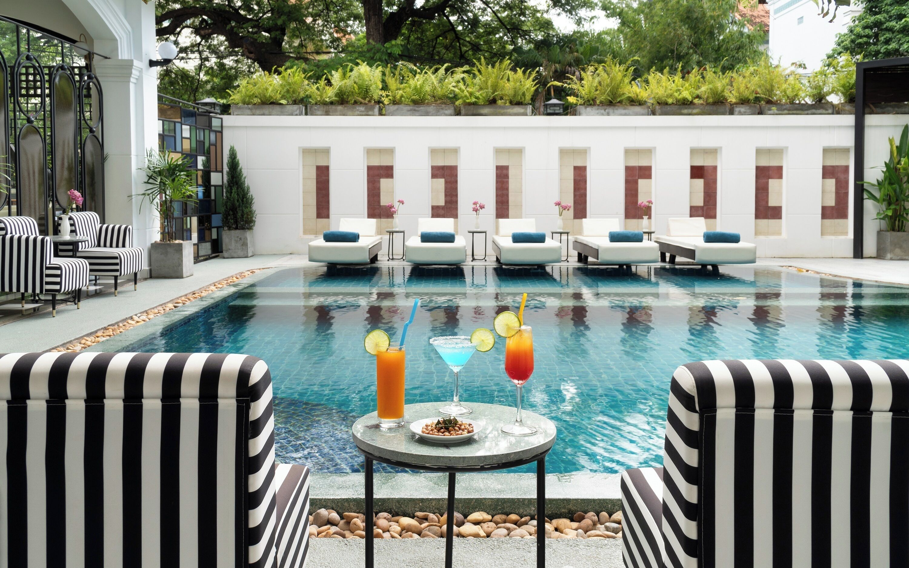 A pool area with lounge chairs, a table with drinks, and a view of a building with windows.