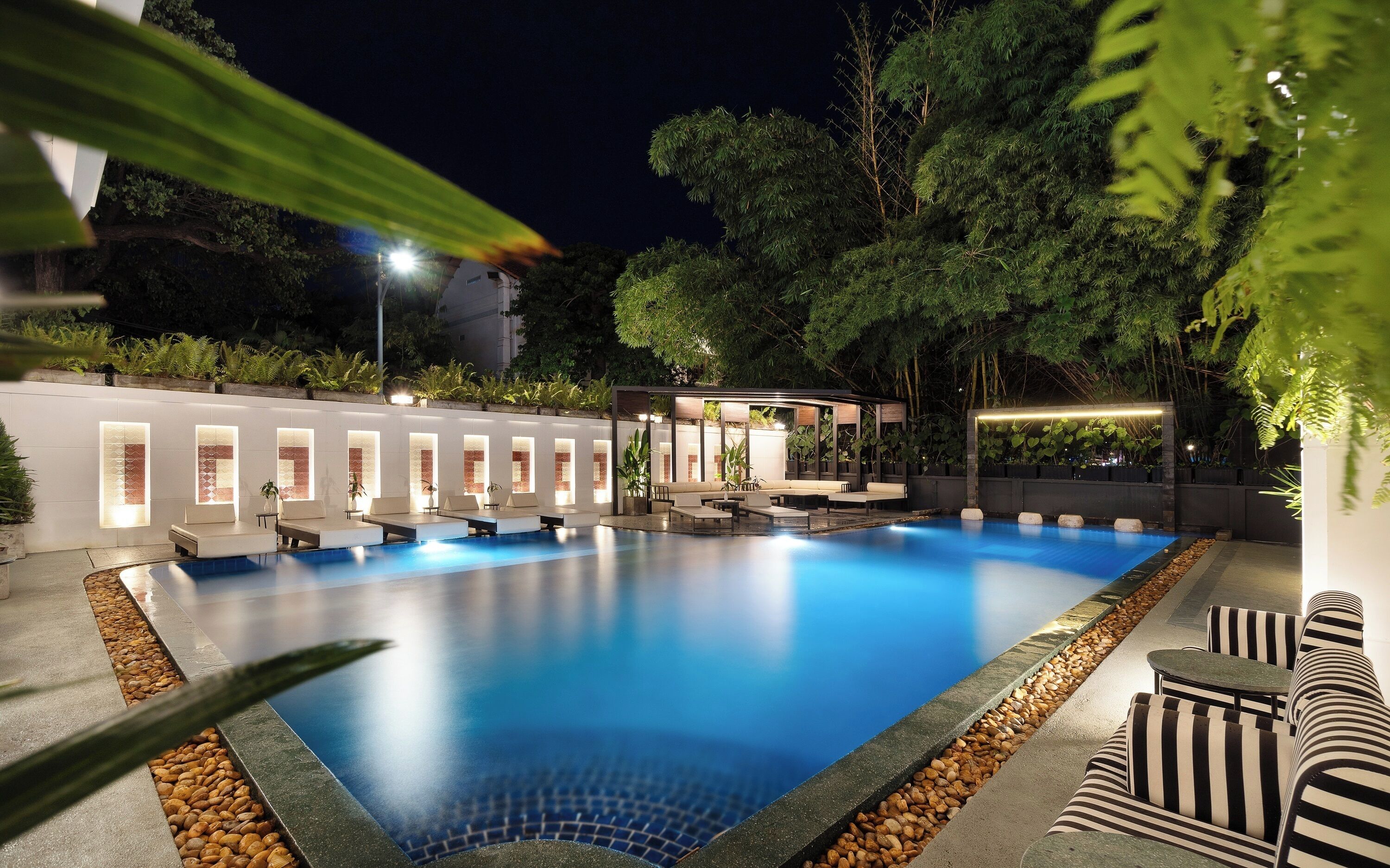 A swimming pool with lounge chairs and a covered seating area, surrounded by greenery and a white wall with windows.