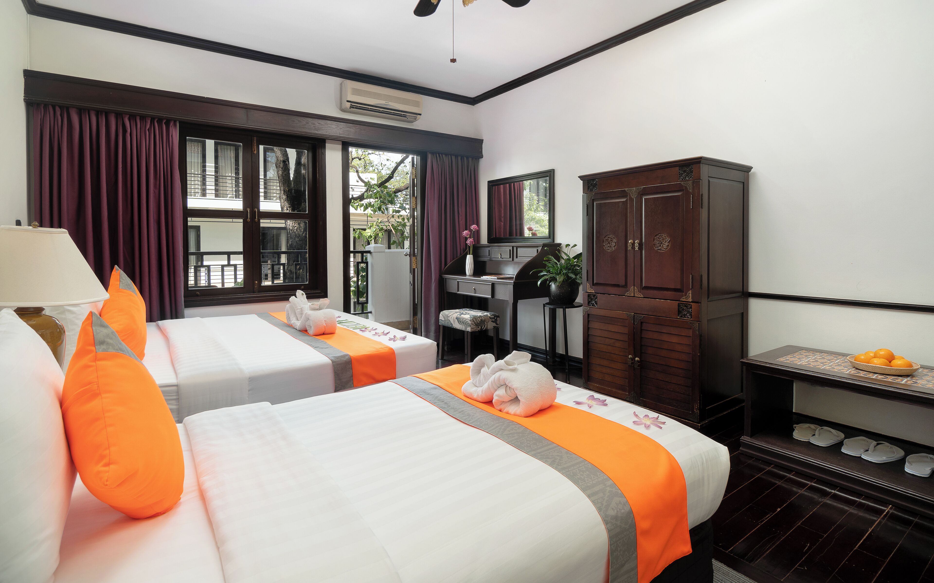 A hotel room with two beds, a desk, a wardrobe, and a small table with oranges.