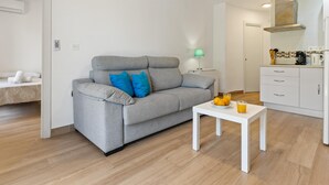 A modern living room with a grey sofa, a small white table, and a kitchen area with white cabinets and a stove. - Torre Copenhague - Terrace, Swimming Pool, Bechfront (Torremolinos)