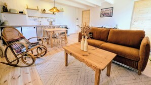 Una sala de estar acogedora con un sofá marrón, un sillón mecedor de madera y una mesa de centro de madera. Hay una zona de cocina con una mesa de comedor y sillas.