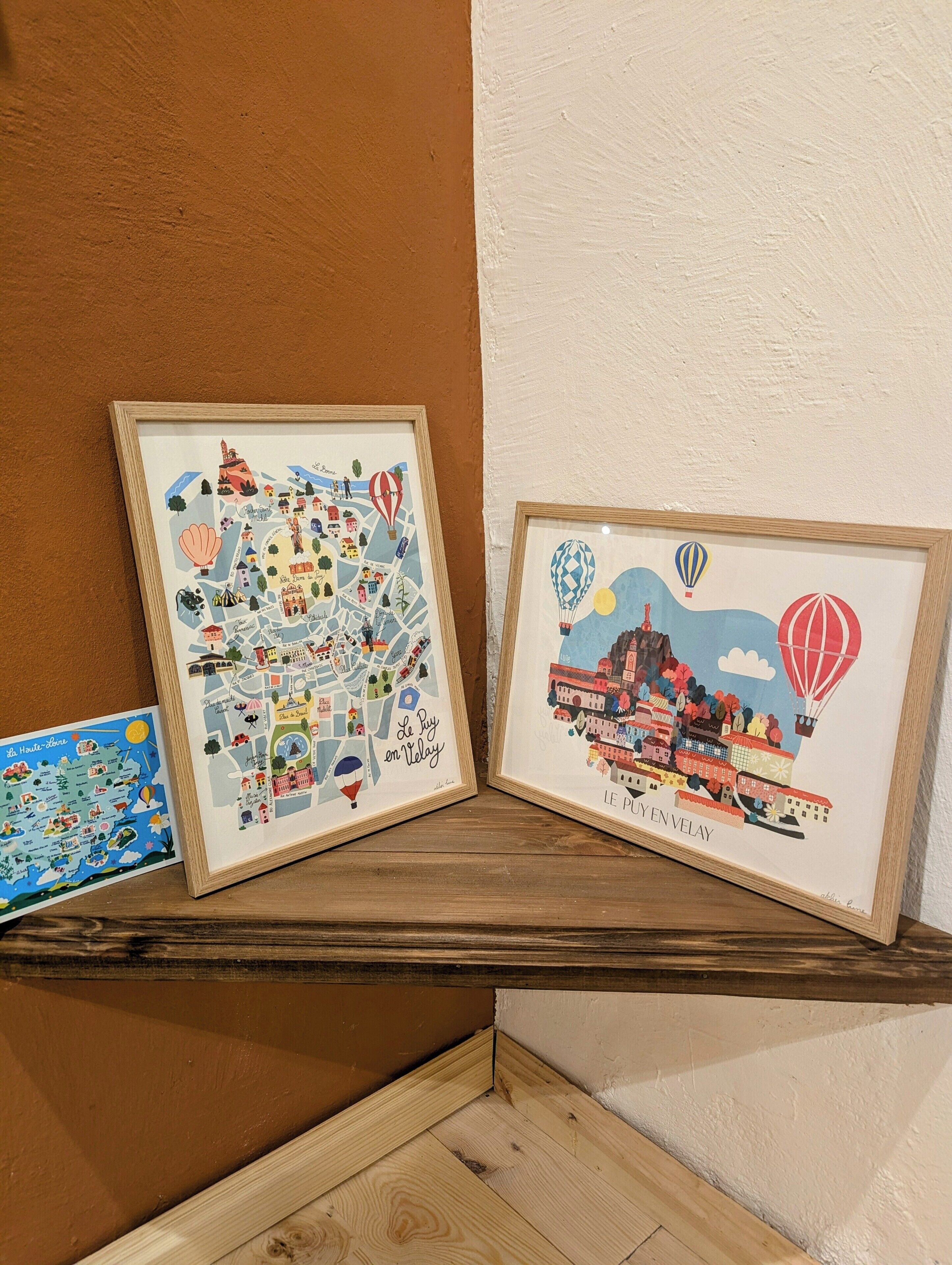 Twee kunstwerken in lijsten op een houten plank tegen een bruine muur.