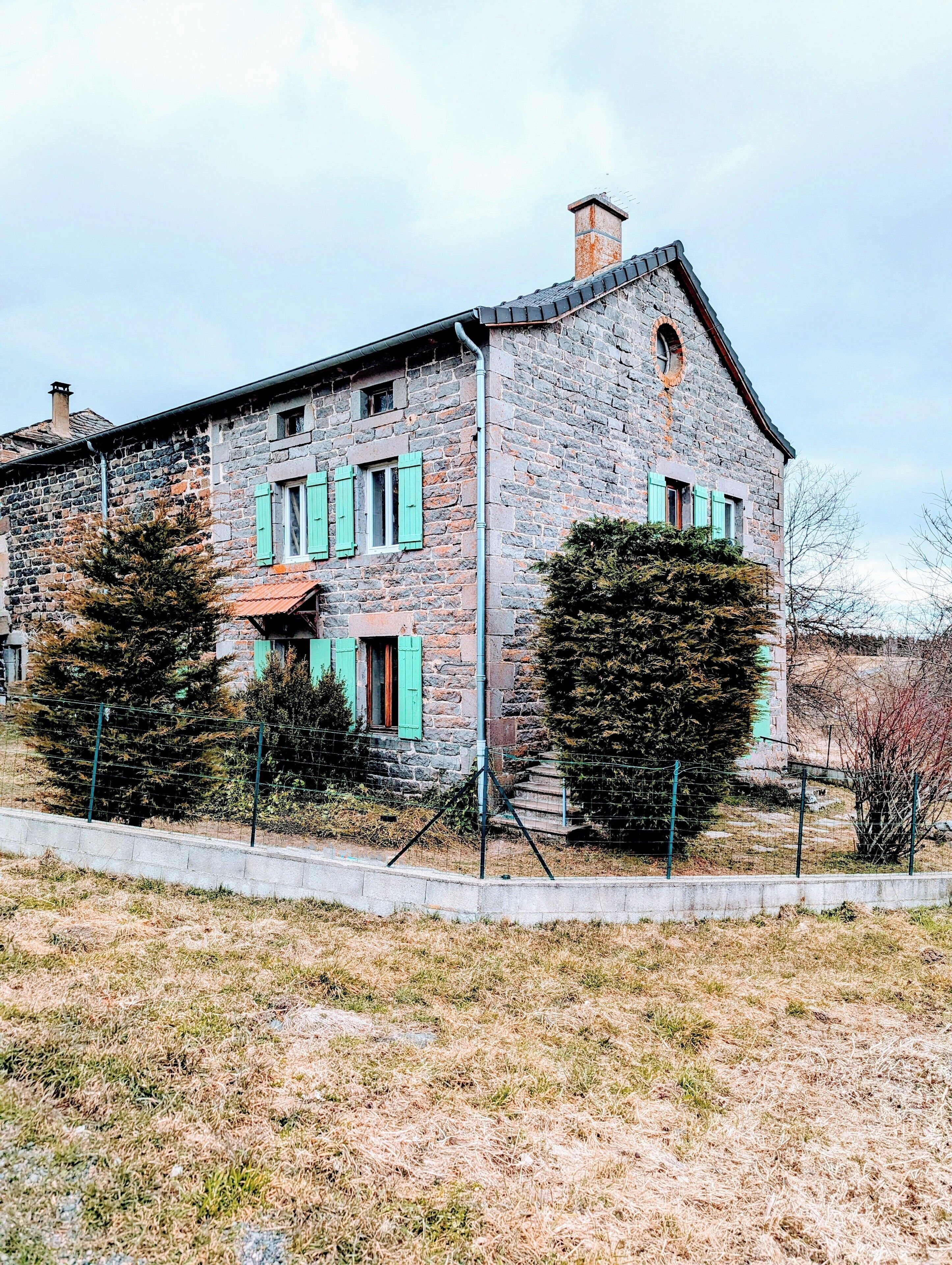 A stone building with a chimney, green shutters, and a fenced yard.