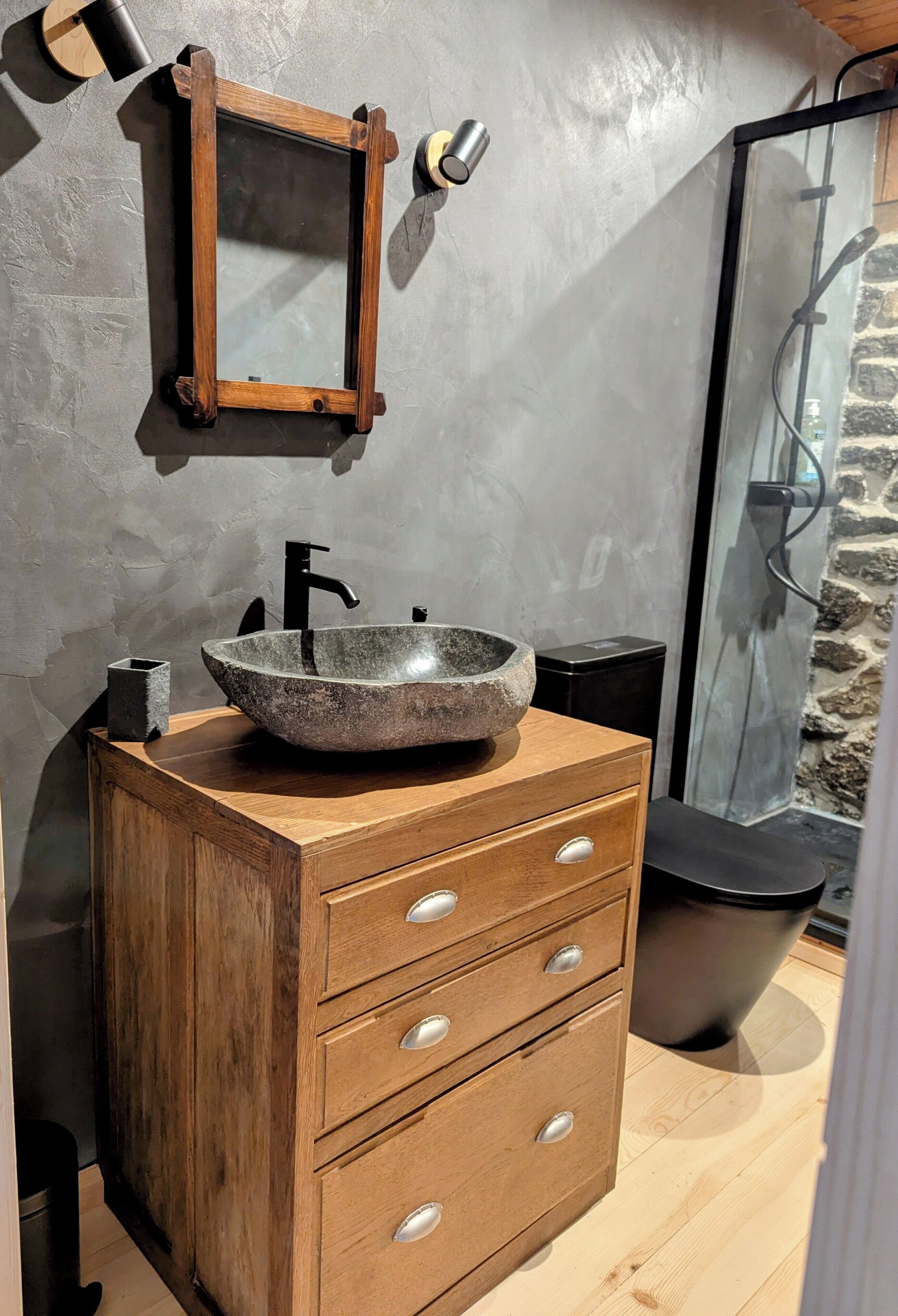 Un baño con tocador de madera, lavamanos de piedra, espejo y zona de ducha.