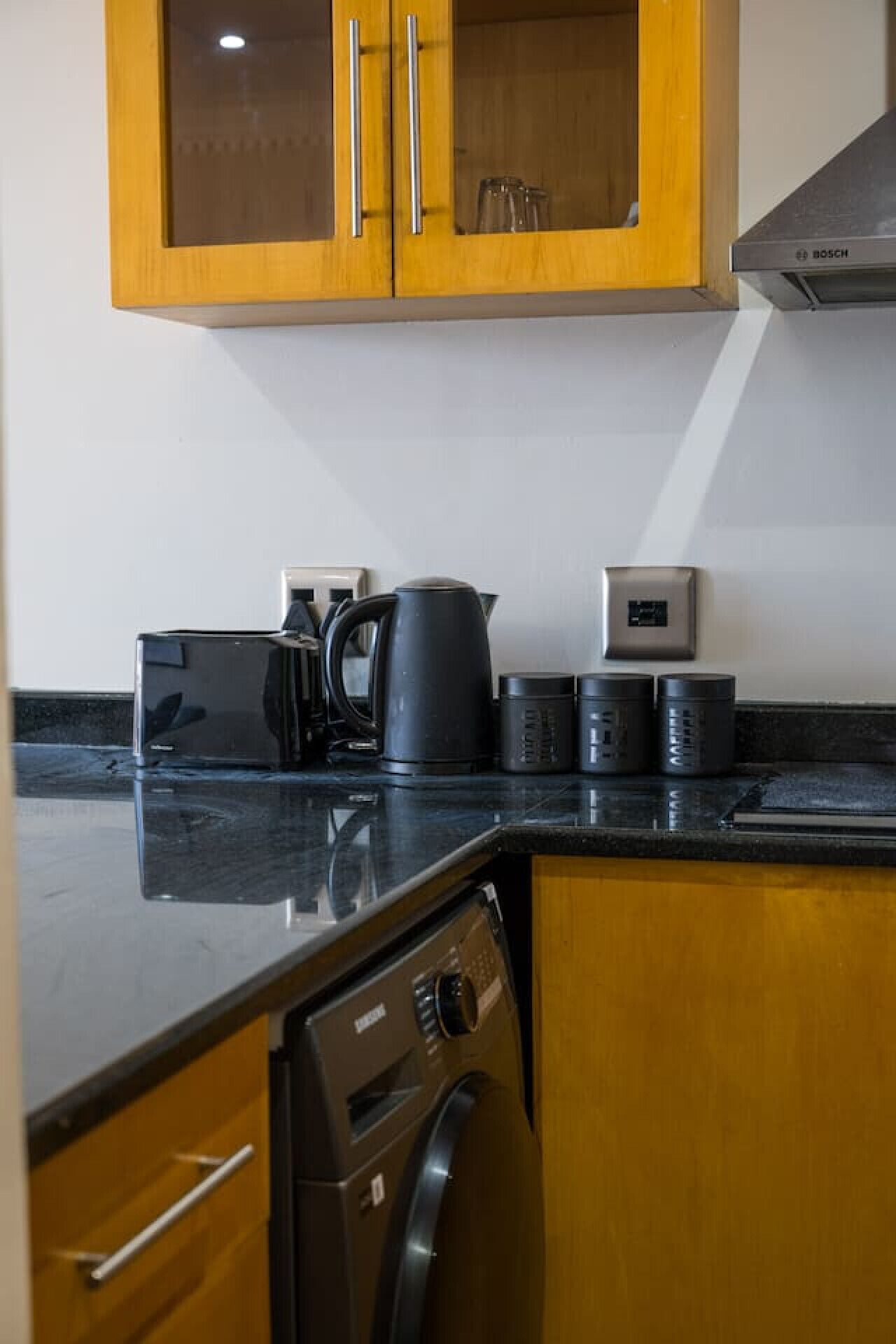 A compact kitchen with wooden cabinets, a Bosch dishwasher, a coffee maker, and a digital scale.