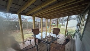 Un patio techado al aire libre con una mesa de vidrio y sillas de mimbre, cubierta de madera y vistas a un jardín.