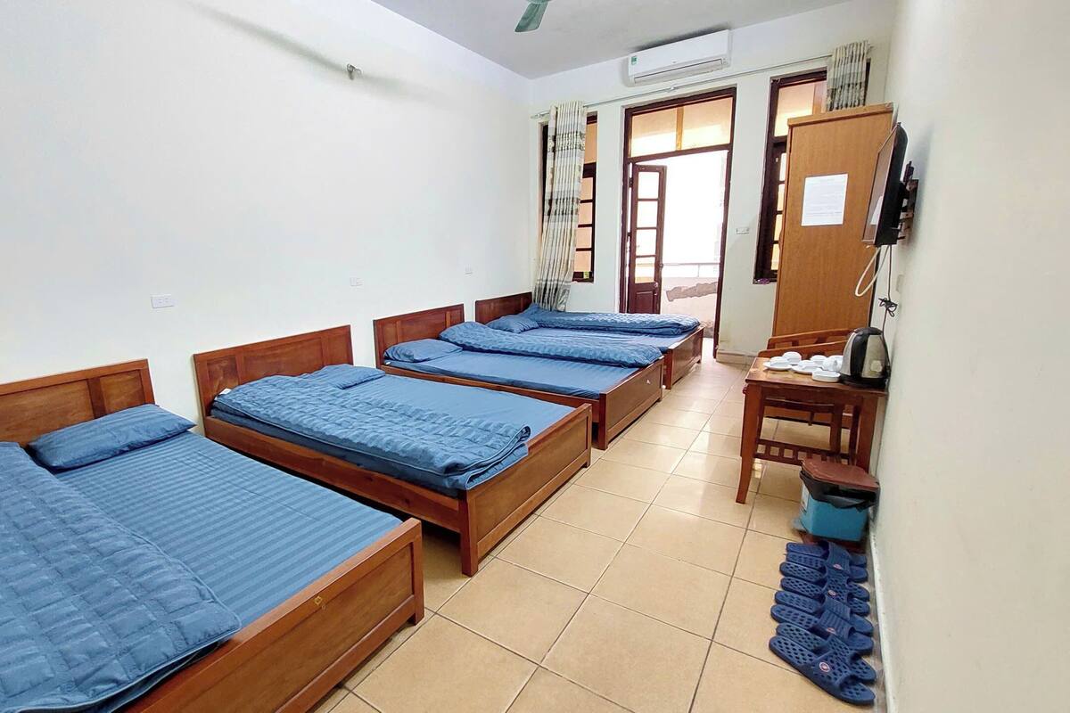 A dormitory room with four wooden beds, a desk with a chair, a small table, and a trash bin.