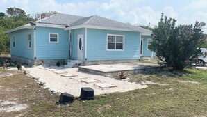 A light blue single-story house with a front porch, a small garden, and a tree with pink flowers. - L&L Cozy Stuido Inn Studio unit Exuma (Exuma)