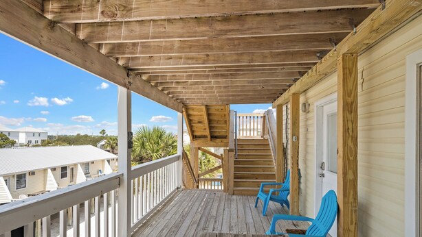 Una terraza de madera con una escalera que lleva a un espacio cubierto, sillas azules y vistas de casas vecinas.
