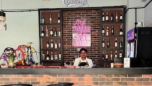 Eine Bar mit einer Neonreklame, einem Barkeeper und einer Ziegelwand.
