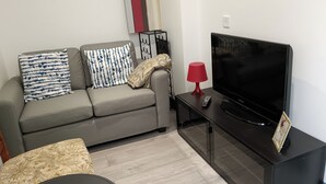 Ein modernes Wohnzimmer mit einem grauen Sofa, gemusterten Kissen, einer schwarzen TV-Konsole und einer roten Lampe sowie einem gerahmten Bild.