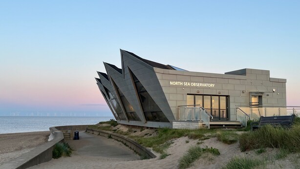 Een modern gebouw met een uniek, hoekig ontwerp, genaamd 'North Sea Observatory', gelegen aan het strand met uitzicht op zee en windmolens op de achtergrond.