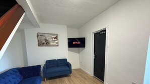 Ein Wohnzimmer mit einem blauen Sofa, einem kleineren blauen Sessel, einem an der Wand befestigten Flachbildfernseher und einer Tür rechts.
