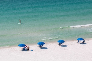A beach with clear turquoise water, a person surfing, and several blue umbrellas and beach chairs.