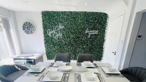 A modern dining area with a central island table set for four, surrounded by dark grey chairs. A green leafy wall with 'Good Vibes Only' and 'be our guest' decor is behind the table. - Perfect 3 bedroom home - Contractors & Families (Wolverhampton)