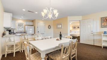 A dining area with a white table and chairs, a chandelier, and a kitchen in the background.