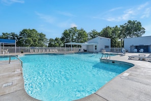 Une piscine avec un bâtiment blanc en arrière-plan, un espace de repos couvert et une zone délimitée par une clôture, aménagée avec des chaises longues.