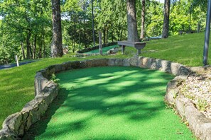 Un green d’entraînement recouvert de gazon synthétique vert, entouré de murs en pierre, avec un banc en bois et un jardin en pierre en arrière-plan.