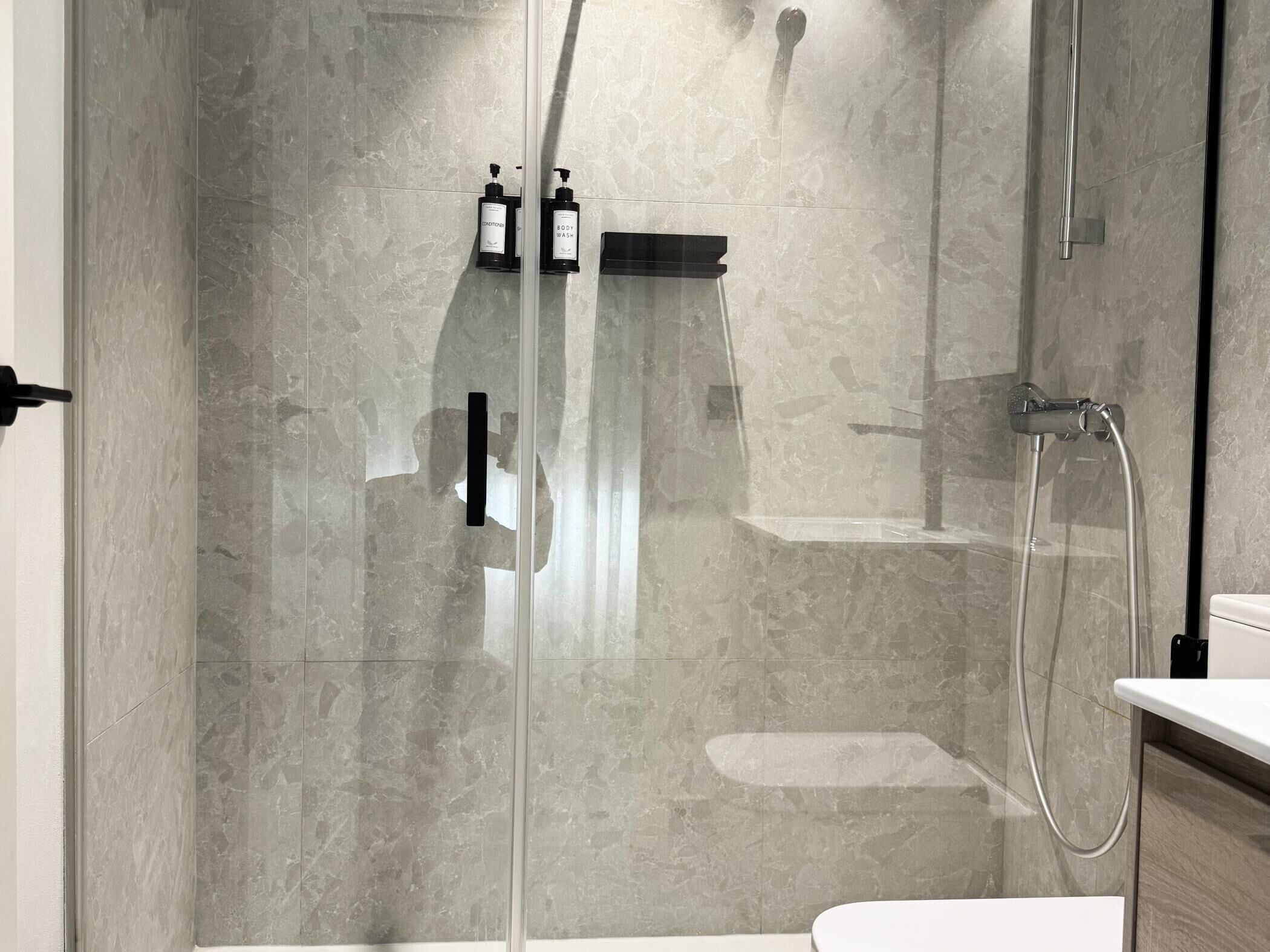 A modern shower with a glass door, a handheld showerhead, and two bottles of shampoo and conditioner on the wall.