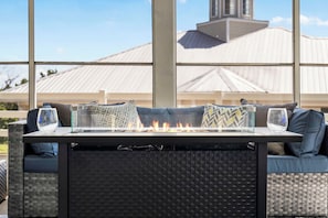Una mesa con fogón, cojines y vasos, con un edificio y un ventanal grande de fondo.