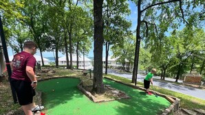 A miniature golf course with a green playing surface, surrounded by trees and a building in the background.