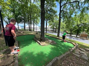 Un campo de mini golf con superficie de juego verde, rodeado de árboles y un edificio al fondo.