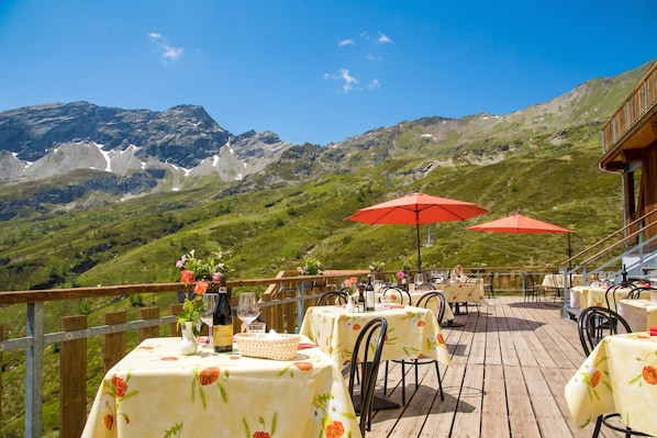 A terrace with tables and chairs, a wooden deck, and a mountainous backdrop.