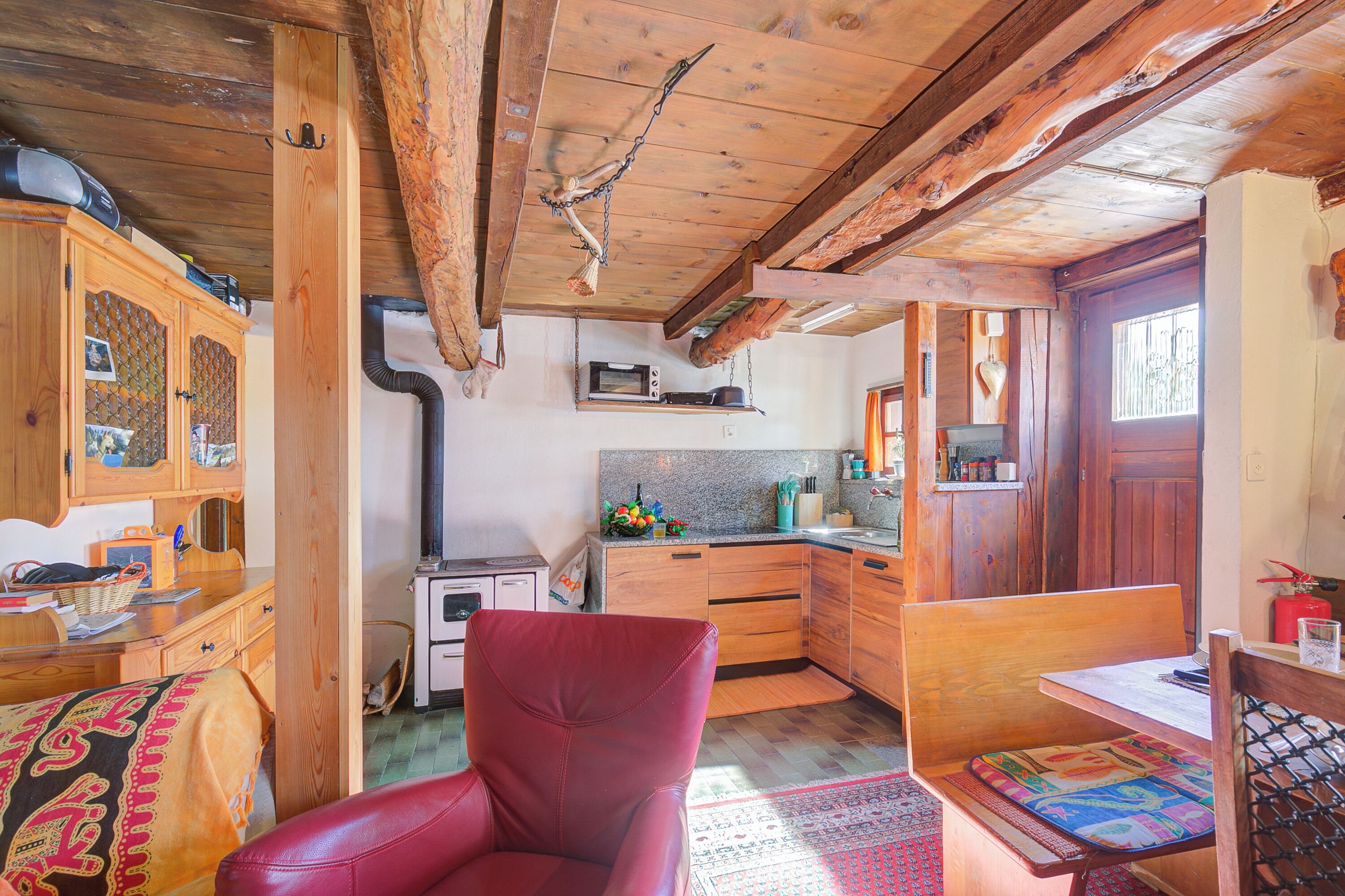 A rustic kitchen with wooden beams, a red leather chair, and a dining table with a patterned table runner.